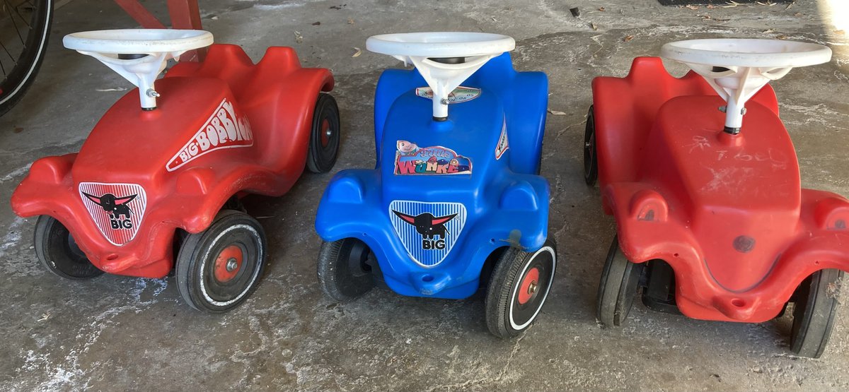 Auf dem Bild sind drei Bobbycars zu sehen. Zwei sind rot, das in der Mitte ist blau. Alle haben ein auffallend weißes Lenkrad.