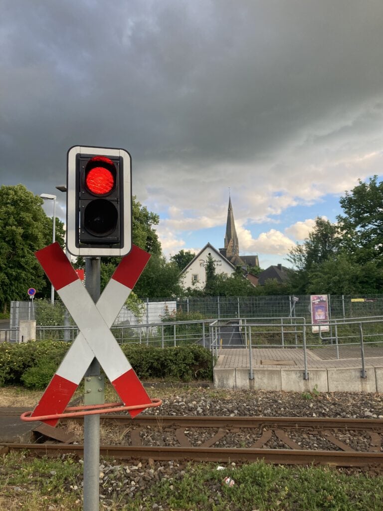 Das Bilde zeigt eine Bake am Bahnübergang in Oesede mit roter Ampel und dem Kirchturm im HIntergrund